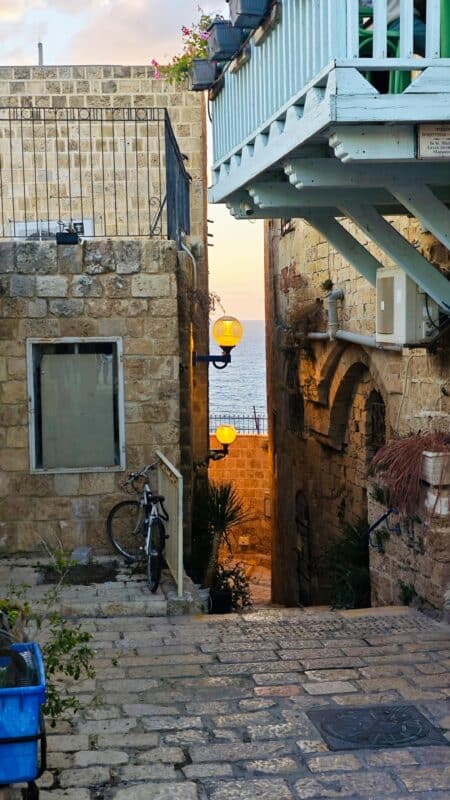 סמטת אבן ביפו העתיקה עם מדרגות, פנסים צהובים ומרפסת עץ תכולה, והים בקצה